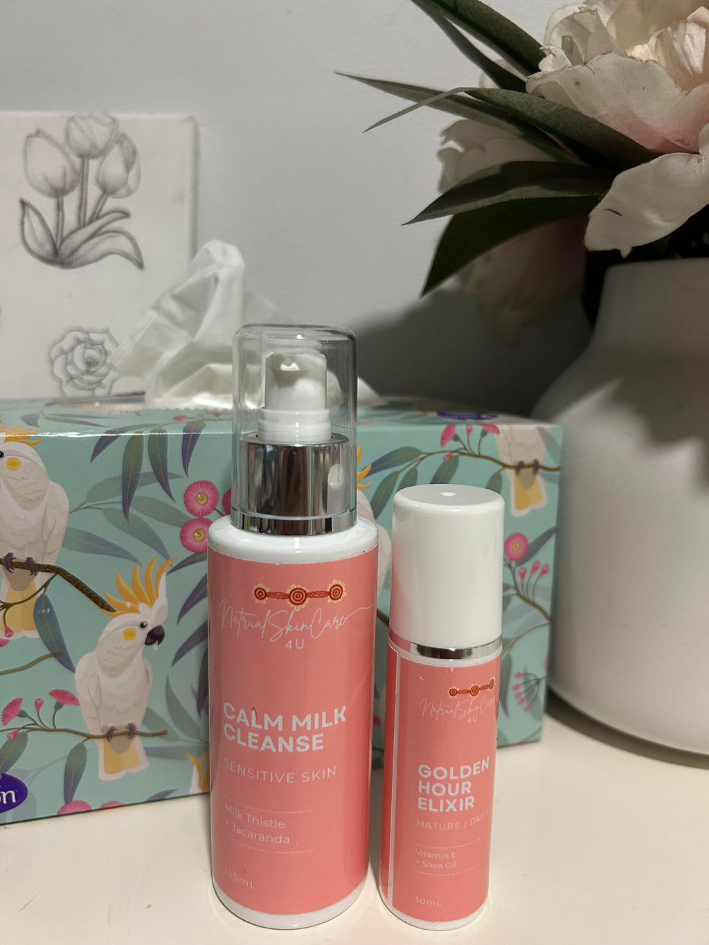 Two skincare products on a surface with a decorative box and flowers in the background