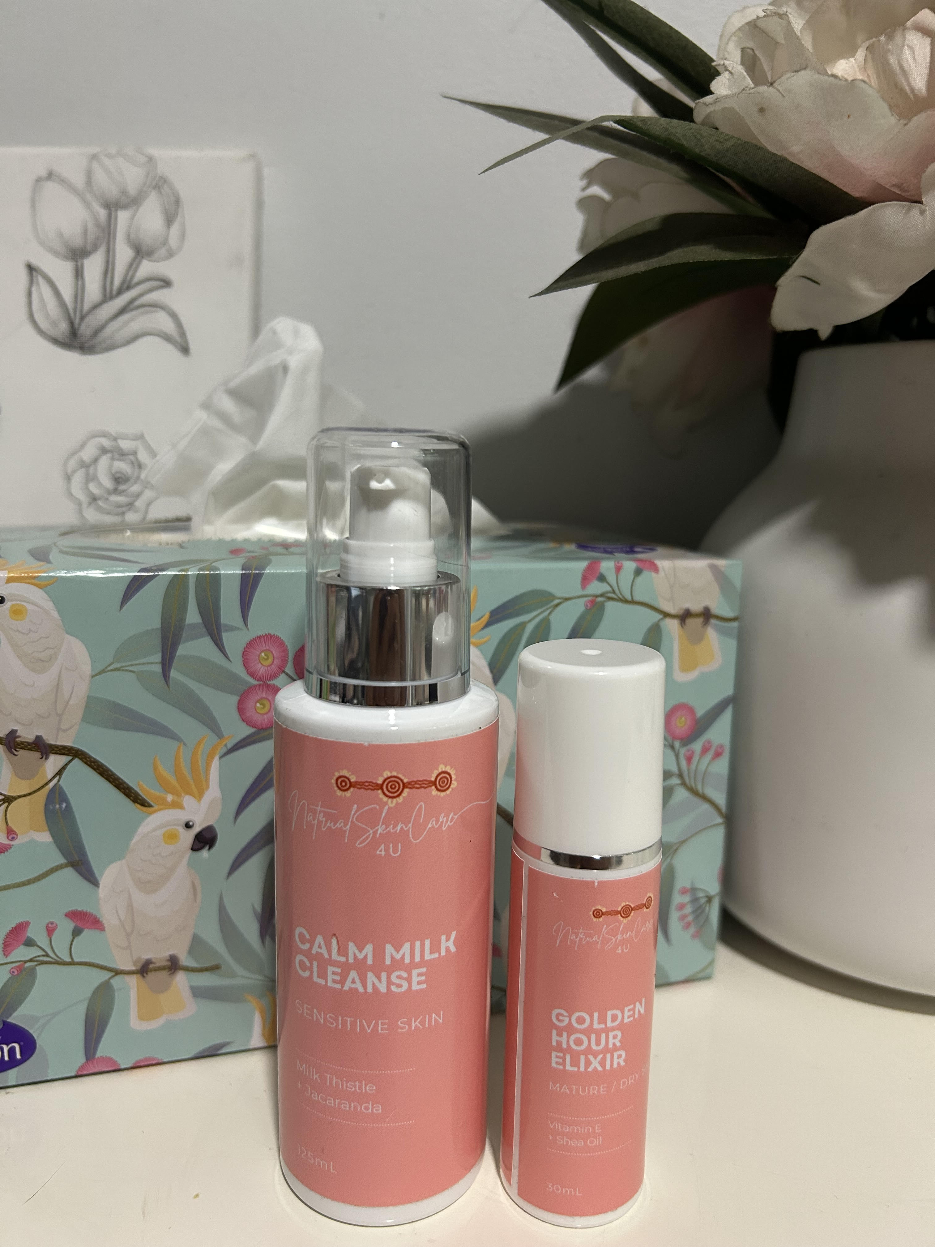 Two skincare products on a surface with a decorative box and flowers in the background